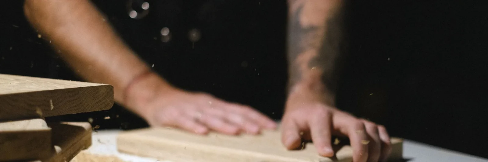 Deux mains à plat sur une planche de bois clair, dans la poussière d'un atelier